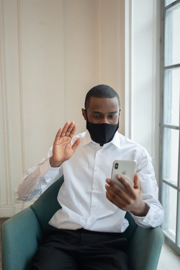Black male executive in textile mask sitting in armchair while showing hello gesture during video chat on cellphone in workspace
